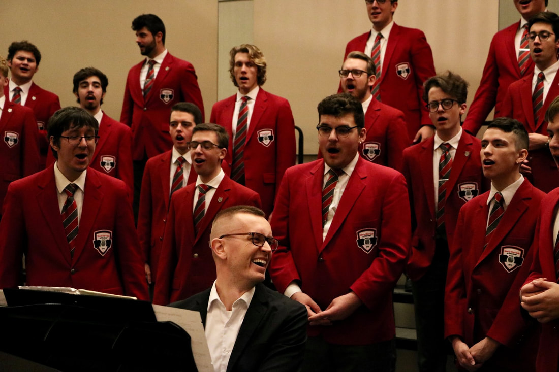 Picture of Mark Wilkinson at the piano looking back at the Ohio State Men's Glee Club with a smile